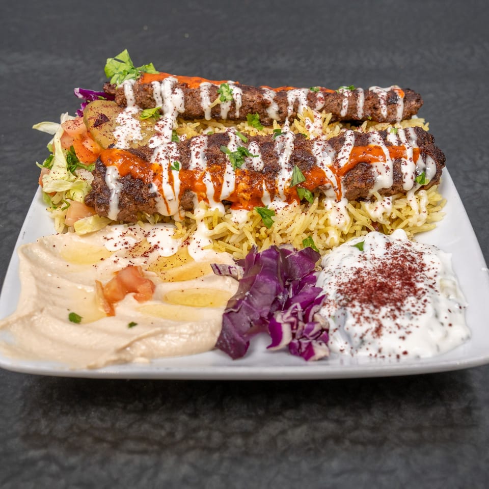 Two kebabs with white and red sauce on rice, salad, hummus, and yogurt dip.